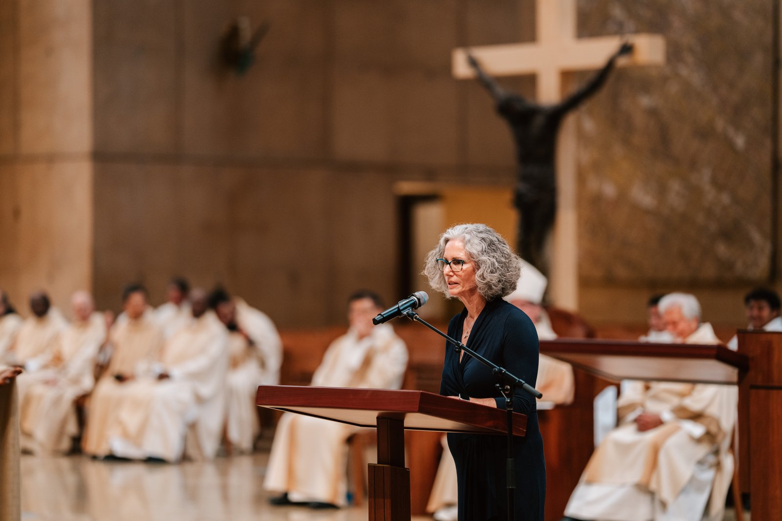 A woman speaks at a podium with a cross of Jesus Christ and priests behind her.