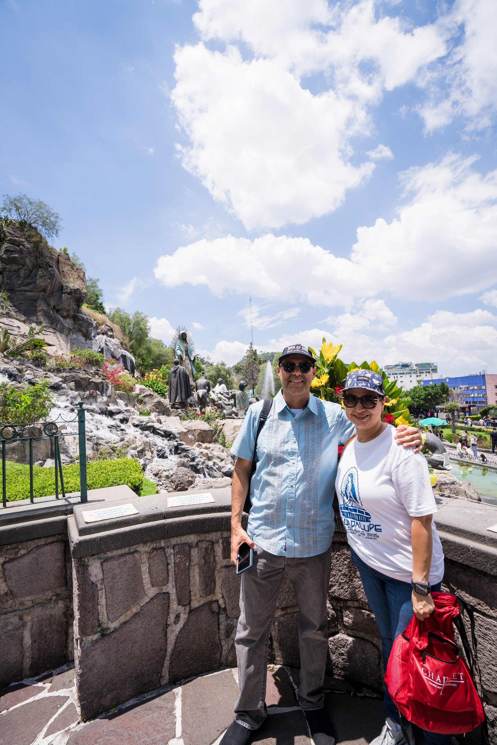 Smiling man and woman pose together outdoors near a rocky garden with statues under a blue sky.