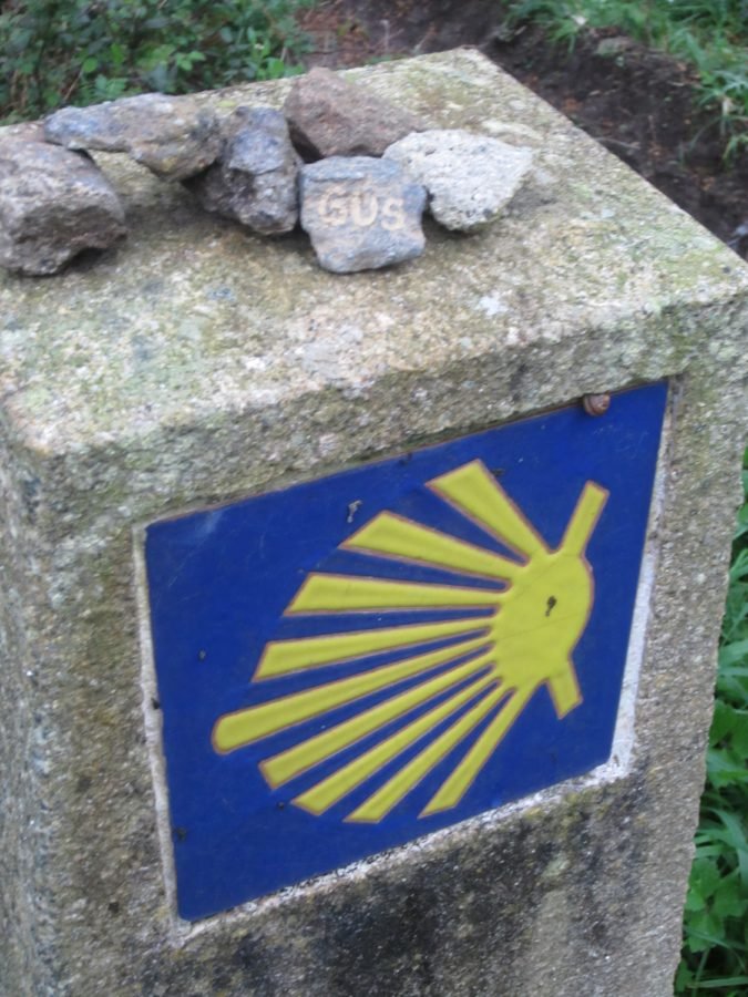 Stone milestone with a blue plaque showing a yellow scallop shell, a Camino de Santiago marker on a mossy concrete post.
