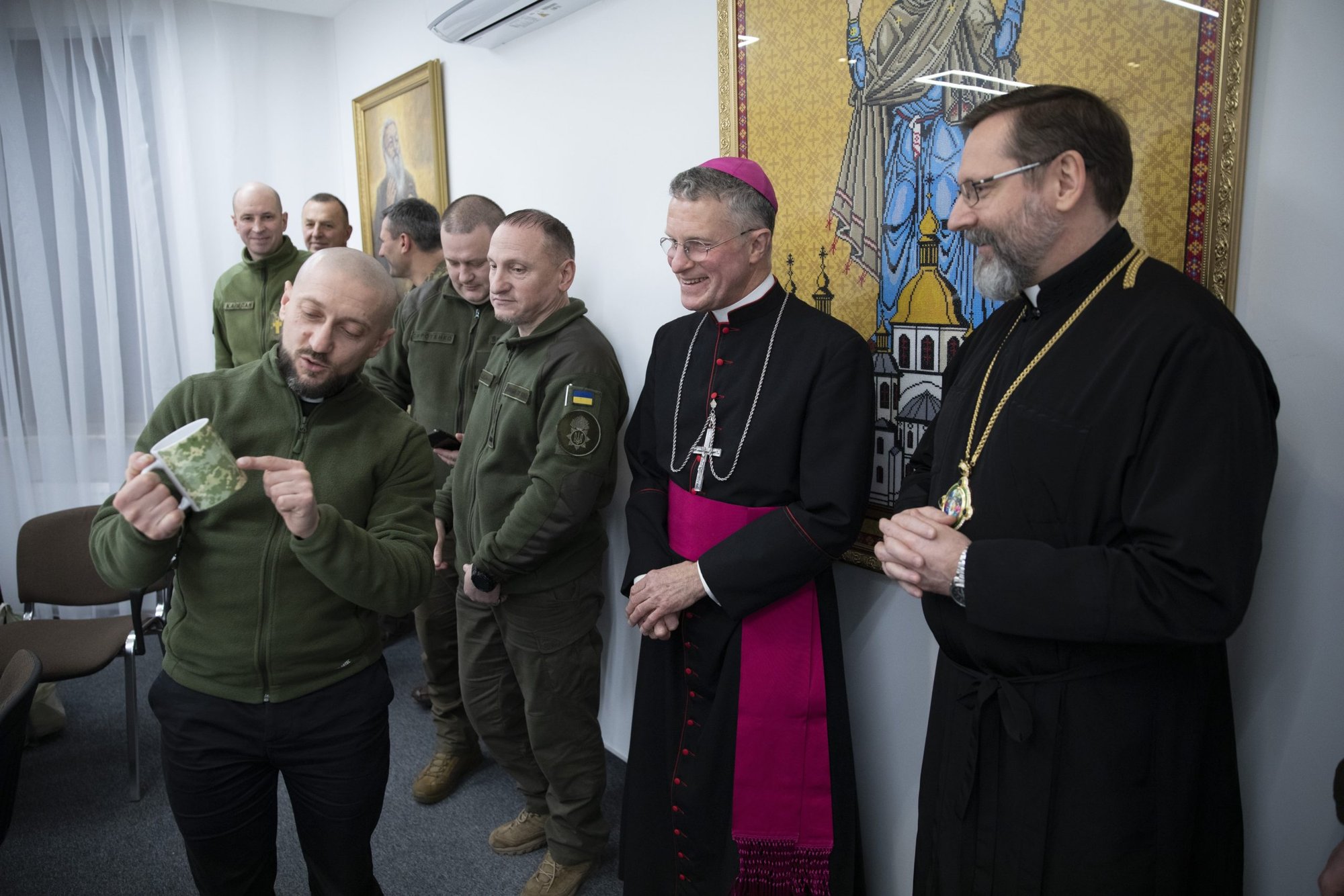 El padre jesuita Andriy Zelinskyy, coordinador de capellanes militares de la Iglesia católica ucraniana, muestra una taza de camuflaje al arzobispo Timothy P. Broglio, jefe de la archidiócesis estadounidense para los servicios militares, durante una reunión en Kiev, Ucrania, el 29 de diciembre de 2022. El arzobispo Sviatoslav Shevchuk de Kyiv-Halych, a la derecha, observa. (CNS /Oleksandr Savransky, cortesía de la Iglesia Católica Ucraniana)