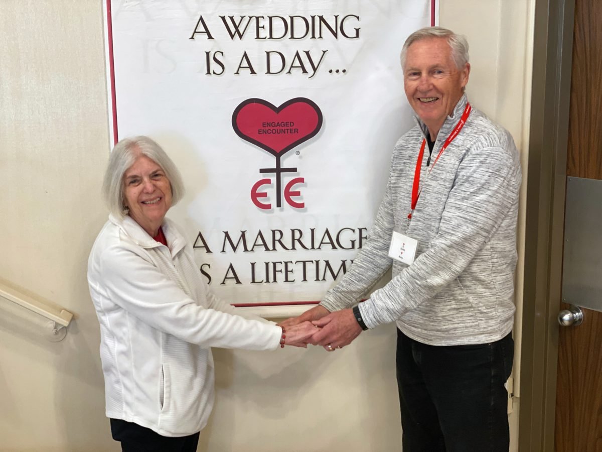 John y Amy Sabol, voluntarios en los retiros de Catholic Engaged Encounters, posan con su foto de boda cerca de un cartel con los temas del fin de semana. (Theresa Cisneros)
