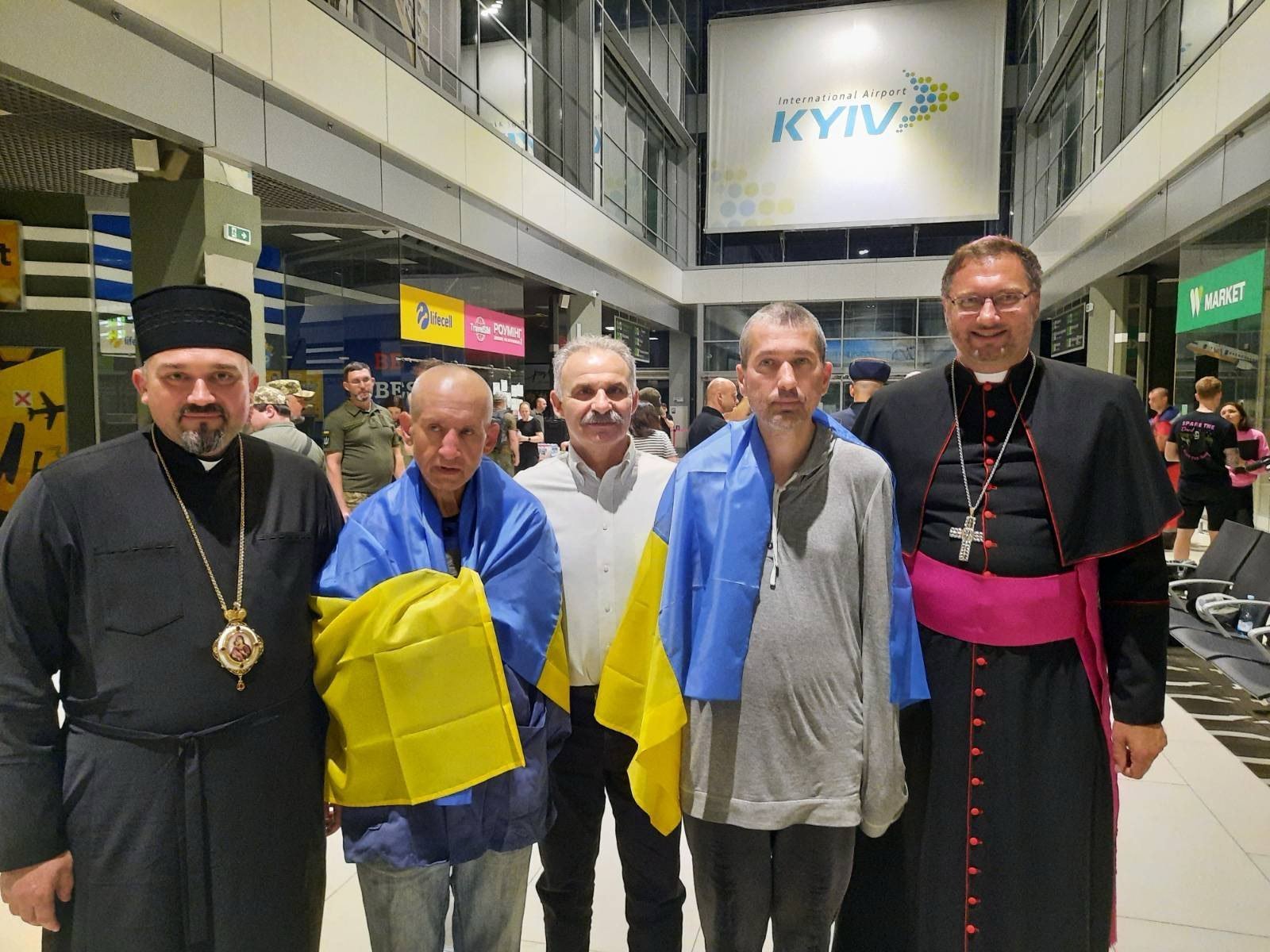 Ataviados con banderas ucranianas, los Padres Redentoristas Bohdan Geleta, a la izquierda, e Ivan Levytsky, a la derecha, posan para una foto en el aeropuerto de Kiev, Ucrania, el 29 de junio de 2024. En el extremo derecho, el arzobispo Visvaldas Kulbokas, nuncio apostólico en Ucrania. (Foto CNS/cortesía del Departamento de Comunicaciones de la Iglesia Católica Griega Ucraniana)