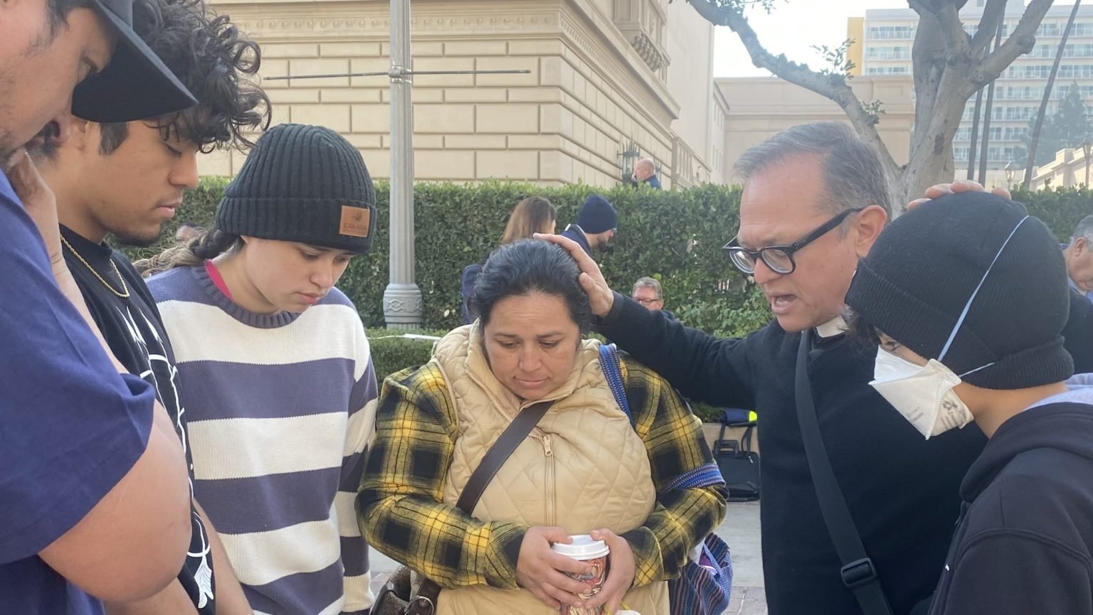 El padre Gilbert Guzmán, párroco de la Iglesia del Sagrado Corazón de Altadena, reza en el refugio de evacuación del Centro de Convenciones de Pasadena con sus feligreses, la familia Posada, cuyo apartamento quedó destruido en el incendio de Eaton. (Natalie Romano)