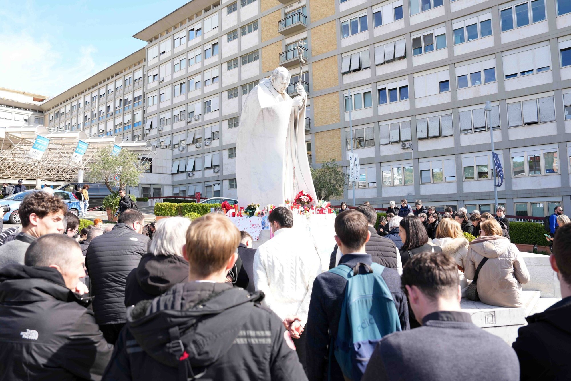 Jóvenes y miembros de la Casa de María, grupos asociados con la Academia Pontificia de la Inmaculada Concepción, y otros fieles rezan alrededor de una estatua de San Juan Pablo II fuera del hospital Gemelli de Roma el 2 de marzo de 2025. El Papa Francisco ha estado hospitalizado desde el 14 de febrero y está recibiendo tratamiento para una doble neumonía. (Foto CNS/Lola Gomez)