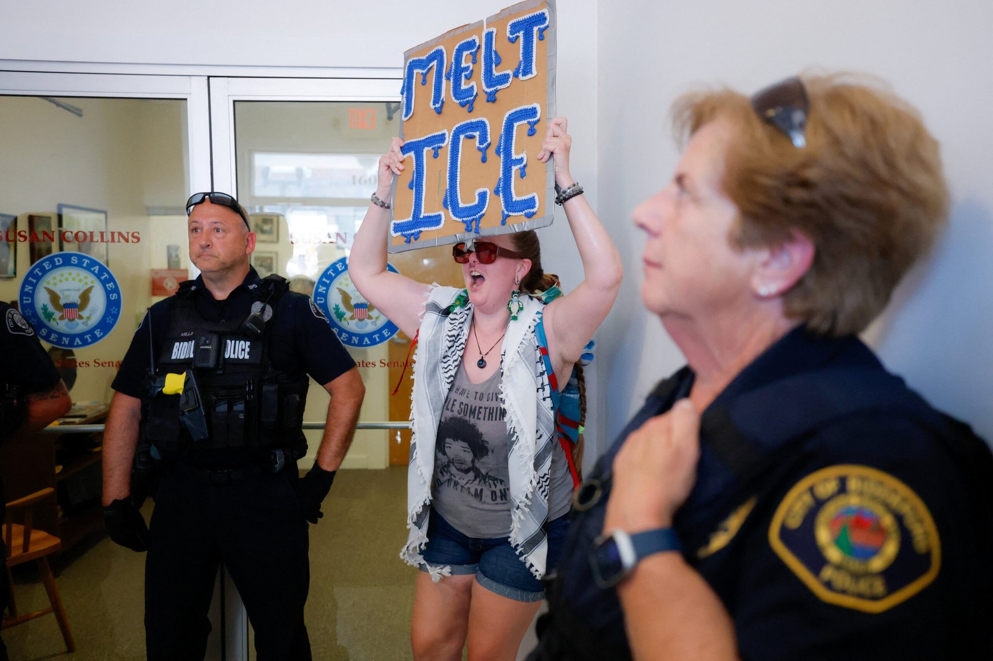 Una mujer sostiene un cartel frente a la oficina de la senadora republicana Susan Collins en Biddeford, Maine, el 13 de julio de 2026, mientras agentes de policía vigilan el lugar y varias personas se congregan para protestar después de que un agente del Servicio de Inmigración y Control de Aduanas (ICE) de Estados Unidos matara a tiros a un automovilista. (Foto de OSV News/CJ Gunther, Reuters)