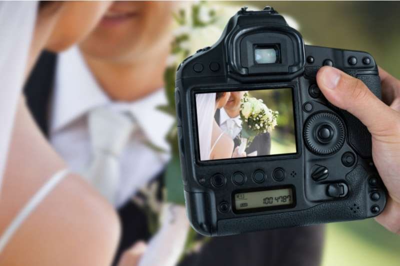Photographer holding camera against couple at wedding. Via Shutterstock