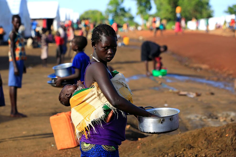 A woman from South Sudan cleans a pot while carrying a child at a refugee camp in Lamwo