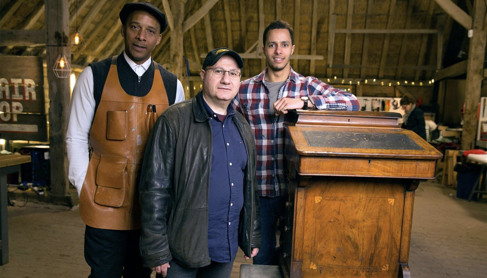 "The Repair Shop" furniture restorers Jay Blades (left) and Will Kirk (right) repaired the highly-prized Davenport desk of Nick Fiaca (center), that had been damaged by burglars. (© Netflix/BBc via Rotten Tomatoes)