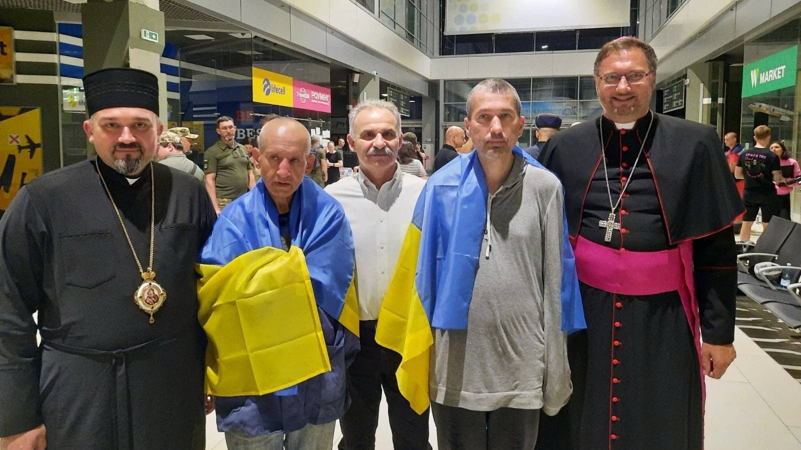 Draped in Ukrainian flags, Redemptorist Fathers Bohdan Geleta, left, and Ivan Levytsky, right, pose for a photo at the Kyiv airport in Ukraine June 29, 2024. To the far right is Archbishop Visvaldas Kulbokas, apostolic nuncio to Ukraine. (CNS photo/Courtesy Ukrainian Greek Catholic Church Communications Department)