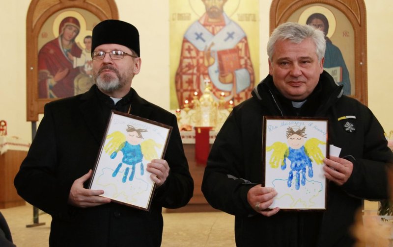 El arzobispo Sviatoslav Shevchuk de Kyiv-Halych, jefe de la Iglesia católica ucraniana de rito oriental, y el cardenal Konrad Krajewski, el limosnero papal, sostienen las huellas de las palmas de las manos creadas por los niños refugiados ucranianos, durante una reunión con los refugiados en Lviv, Ucrania, el 10 de marzo de 2022. (Foto CNS photo/cortesía de la Iglesia católica ucraniana)
