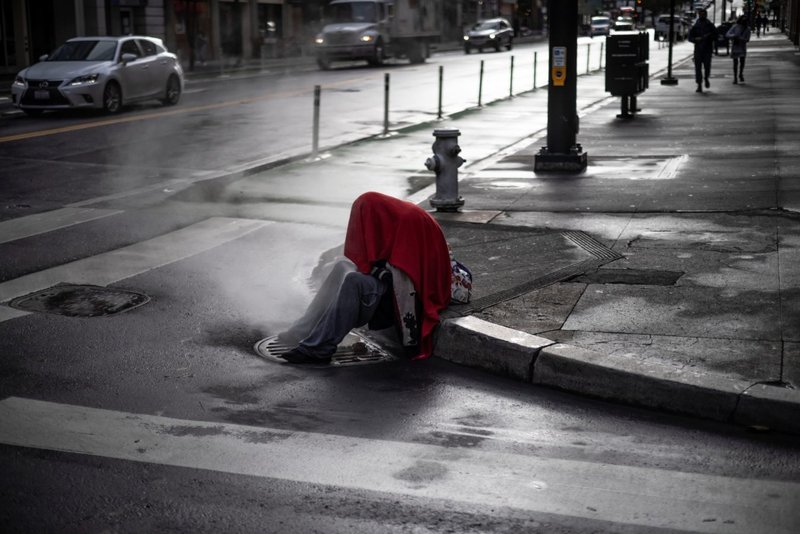 Una persona sin hogar en San Francisco se sienta en una esquina mientras emerge vapor de una rejilla el 22 de octubre de 2021. (CNS/Carlos Barria, Reuters)