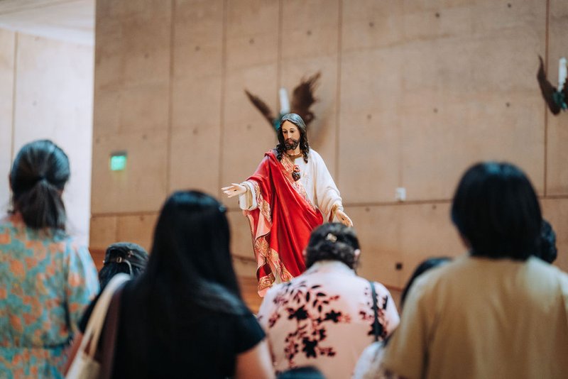 performer dressed as Jesus in white and red robes speaks to a seated audience in a large hall.