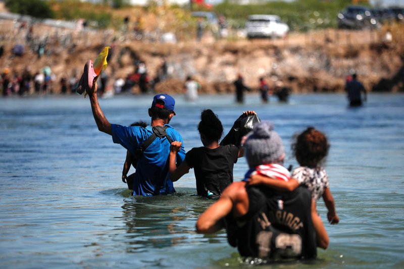 Group of people wearing life jackets wading through water toward a shore, with a rocky embankment and onlookers in the background.