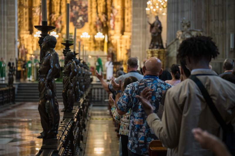 Congregation lined along a railing inside a grand church, hands raised toward a gold-lit altar with statues in the background.