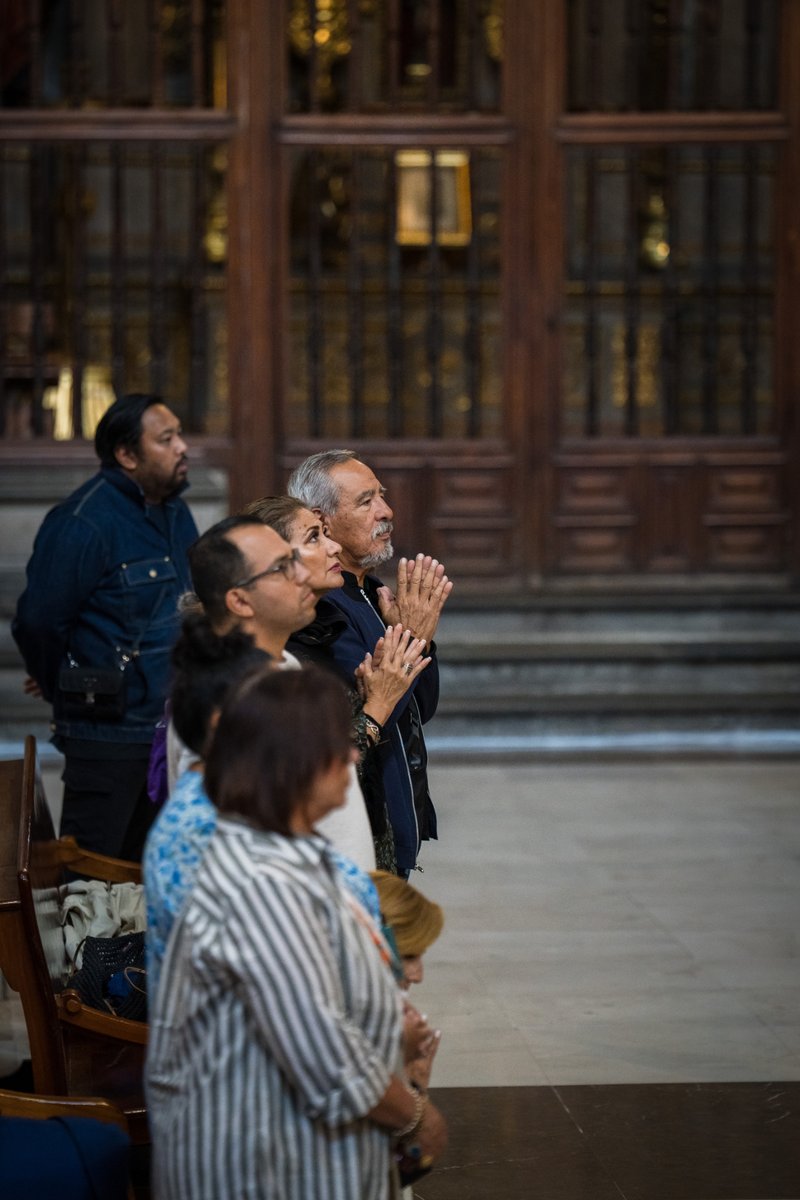 Group of people standing and praying in a church, hands in prayer, with a wooden panelled background and pews nearby
