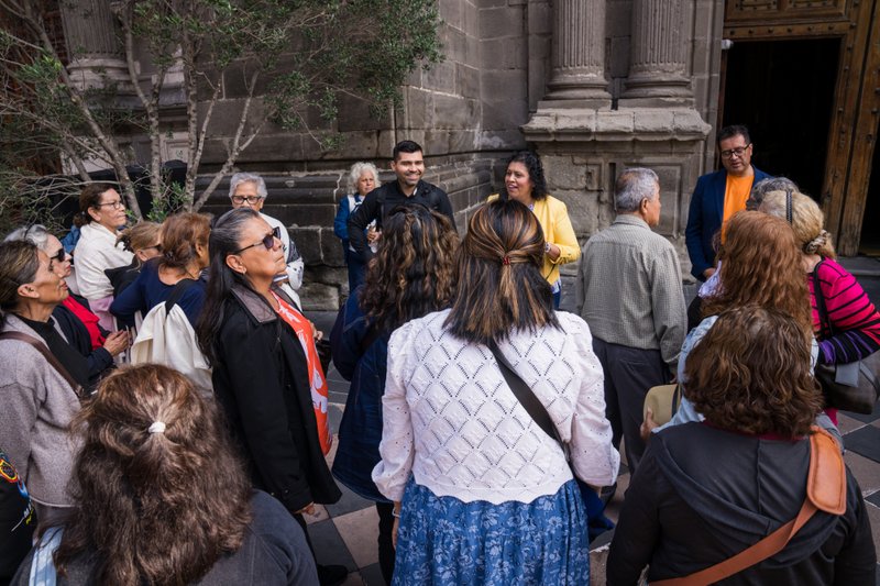 Tour group of adults standing outside a historic stone building with columns, listening to a guide in a yellow jacket.