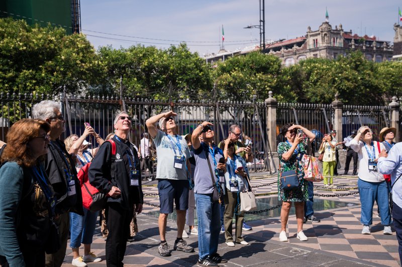 Group of tourists with lanyards and cameras standing behind a decorative fence in an outdoor city square, looking up at something above.