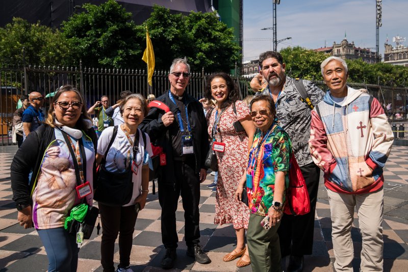 Group of diverse adults with lanyards posing for a photo in a sunny urban plaza, fence and historic buildings behind them.