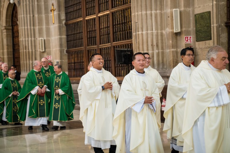 Procession of clergy in green and white liturgical vestments inside a church nave with stone columns and wooden doors in the background.