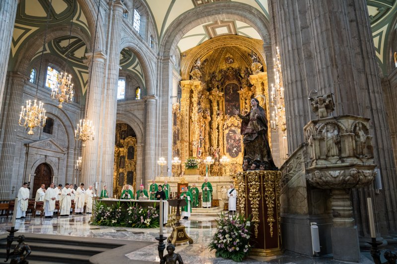 Catholic mass in a grand cathedral with a gold ornate altarpiece, clergy in white and green vestments at the altar, and chandeliers overhead.