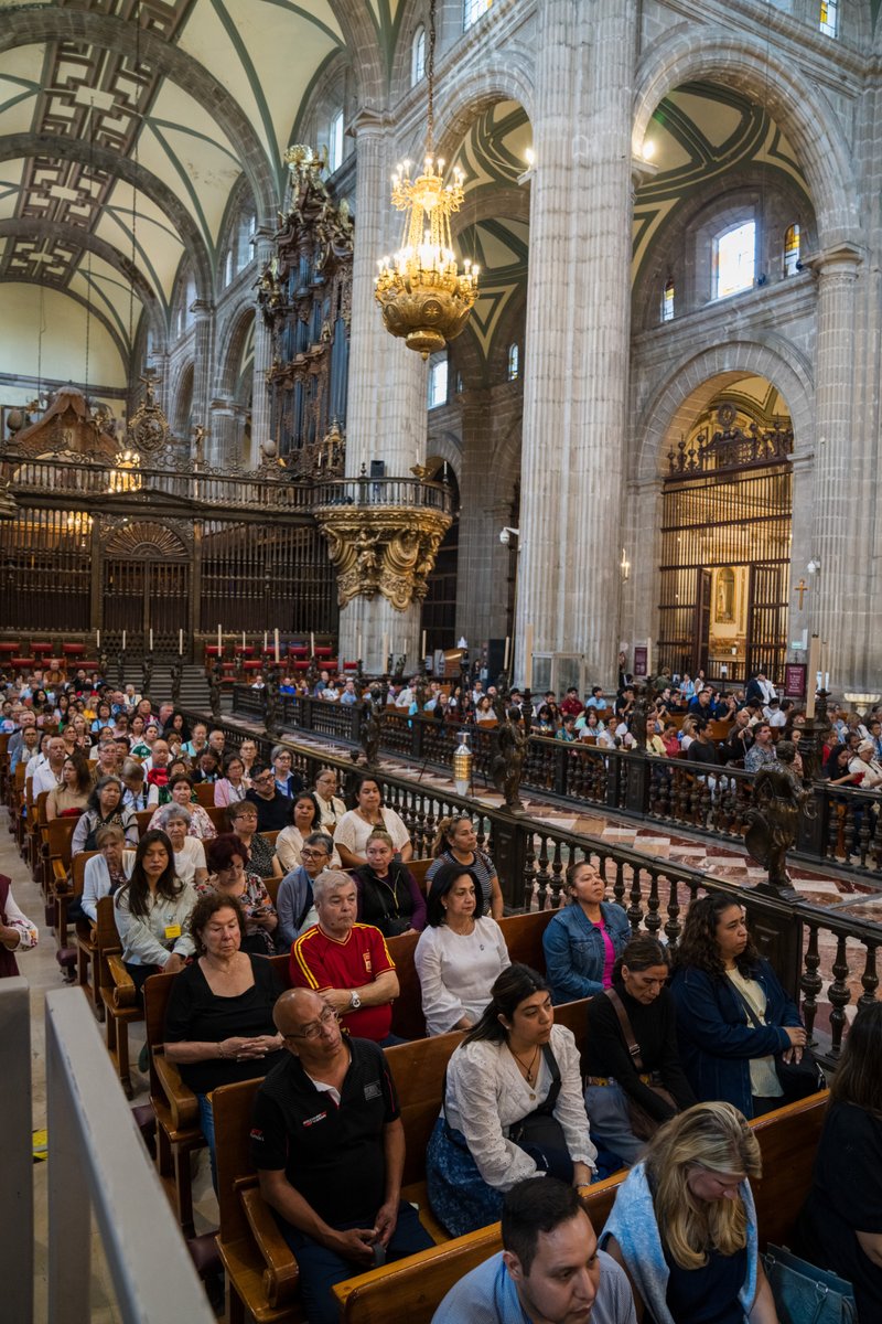 Inside a grand cathedral, a crowded congregation sits in wooden pews beneath towering stone arches and a gold chandelier overhead.