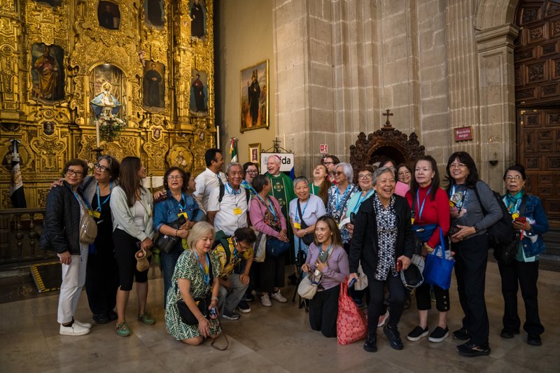 Group of tourists posing for a photo inside a richly gilded church with an ornate gold altarpiece and stone arches in the background, wearing lanyards and smiles.