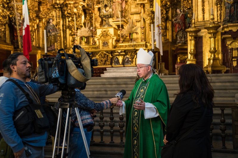 A bishop in green liturgical robes with a white mitre being interviewed by reporters as camera crews film inside a richly gilded church interior.