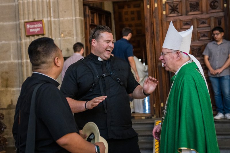 Two men greet each other outside a church doorway: a smiling priest in black clerical attire and a bishop in green vestments with a tall white mitre.