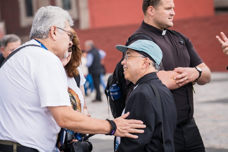 Two men in the foreground shake hands as a photographer looks on, with a priest and others chatting in the background.