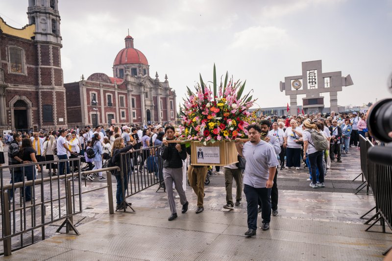 Crowd watches a flower-filled procession float carried through a plaza, with a red-domed historic building in the background.