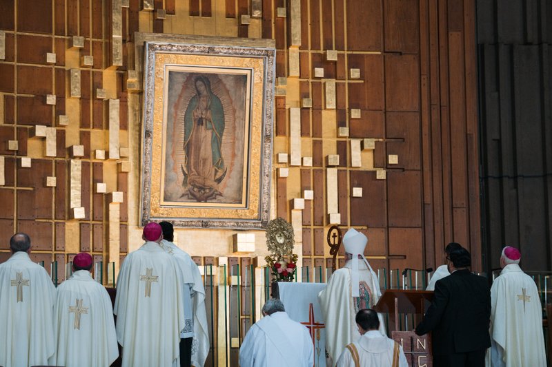 Clergy in white vestments stand before a gold-framed painting of the Virgin Mary inside a church sanctuary.