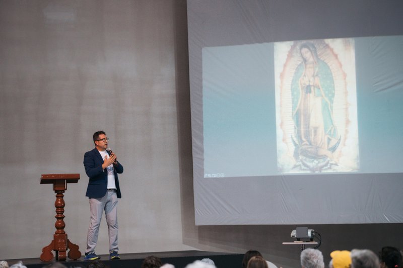 Man in a blue jacket speaks into a microphone on stage beside a wooden podium, with a large projected image of a religious icon on the screen behind him.