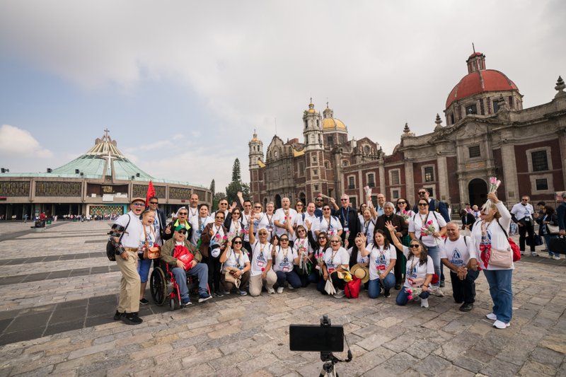 Group of cheerful tourists wearing white shirts posing for a photo in a historic plaza with a grand church featuring yellow domes in the background.