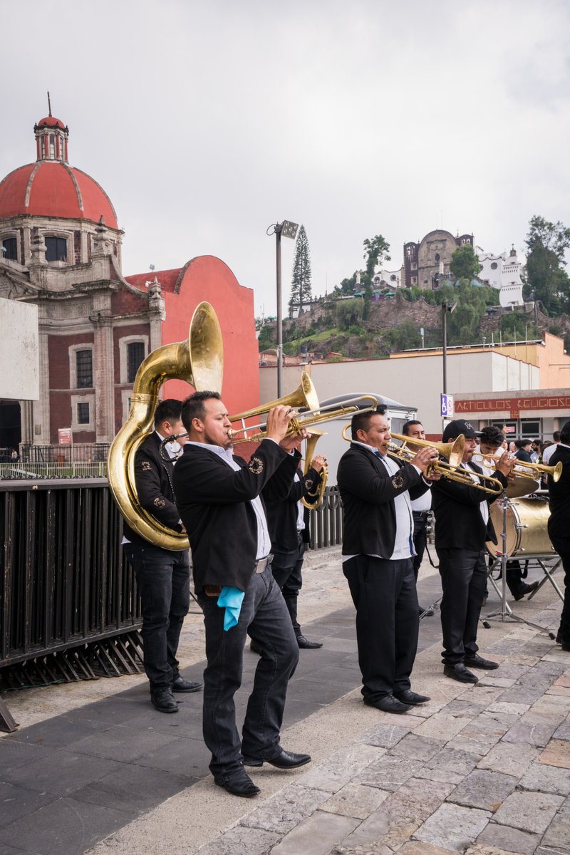 Brass band musicians in black jackets playing trombones on a sidewalk with a red-domed historic building behind them.
