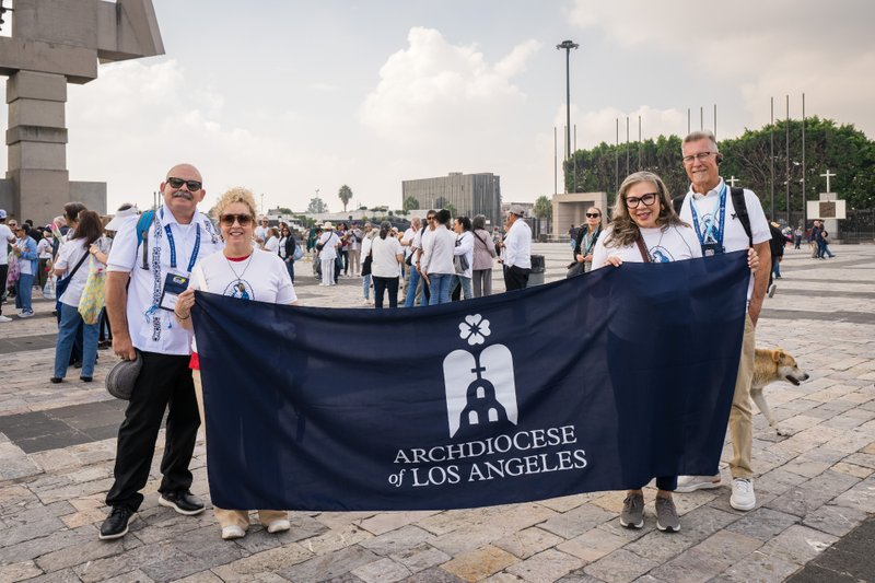 Group of smiling adults holding a dark banner reading Archdiocese of Los Angeles in a sunny plaza with a crowd behind them.
