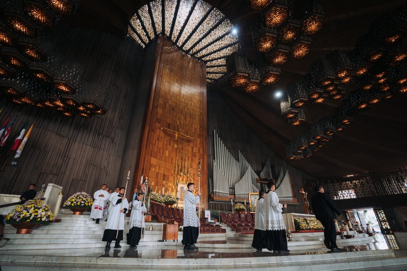 Clergy in white liturgical robes stand on marble steps at a church altar beneath dark wood walls and decorative hanging lights.