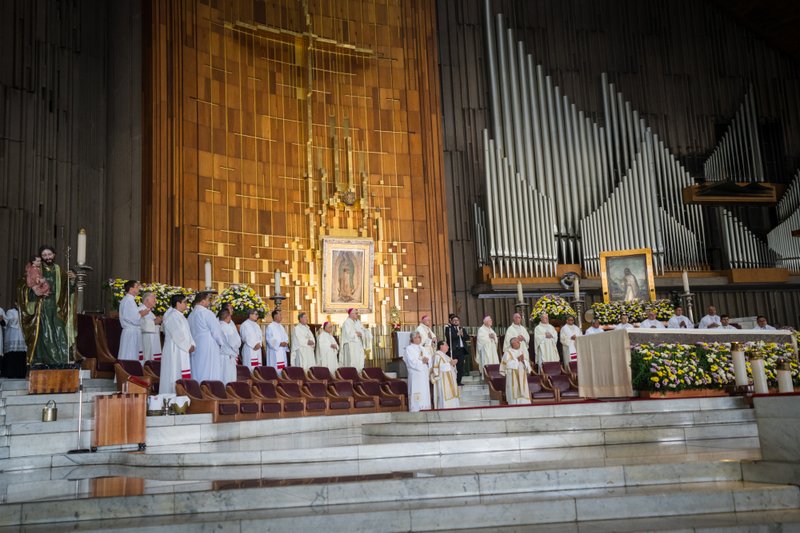 Mass ceremony in a church with clergy in white vestments at the altar, gold panel backdrop and organ pipes in the background.