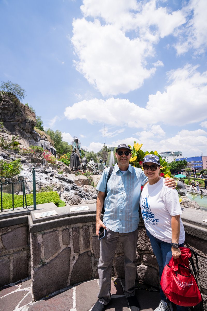 Two people pose together outdoors near a stone wall and statue garden on a sunny day.