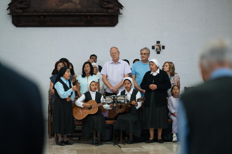 Group of choir members performing, including two guitarists, in a church setting with a white wall backdrop and audience in foreground.
