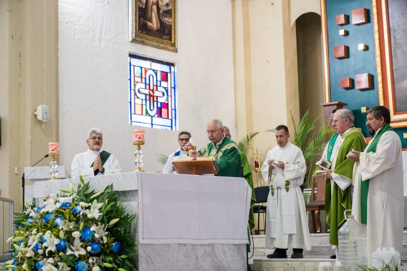 Clergy in green vestments celebrate Mass at an altar with a chalice, candles, and a large floral arrangement inside a church.