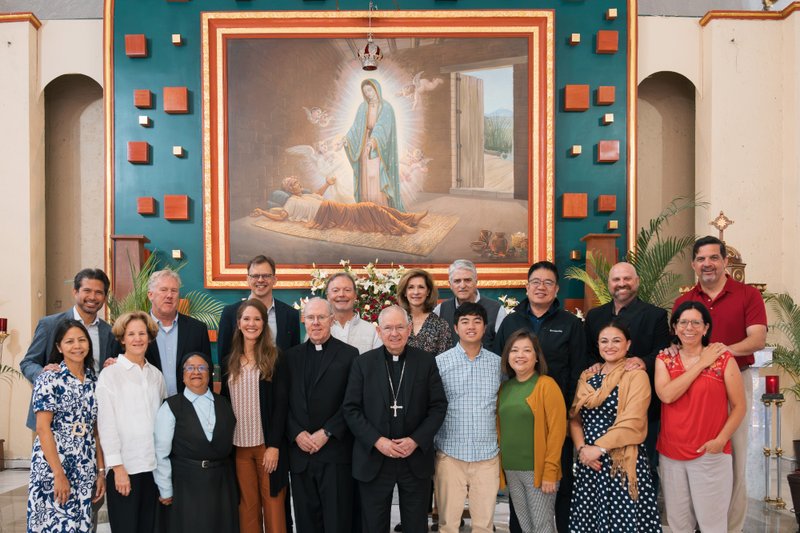 Group of diverse adults posing for a photo in a church in front of a large painting of the Virgin of Guadalupe.