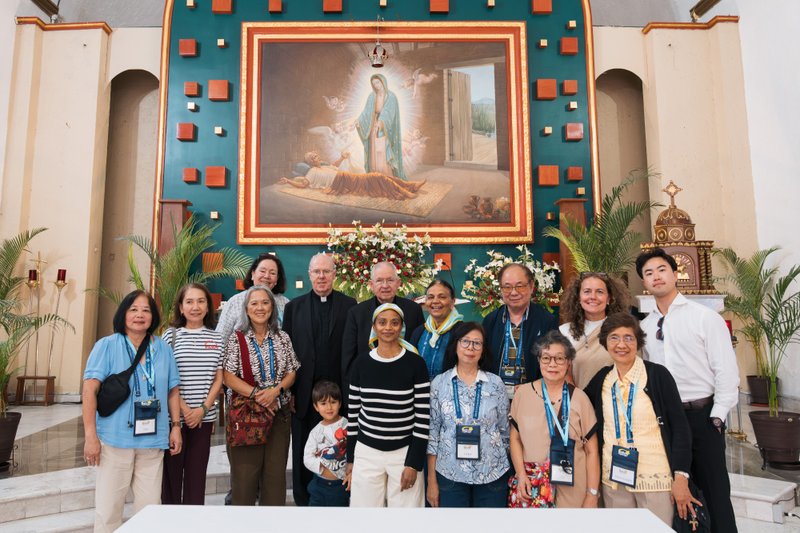 Group of people posing for a photo in a church, in front of a large framed Mary painting and floral altar backdrop.