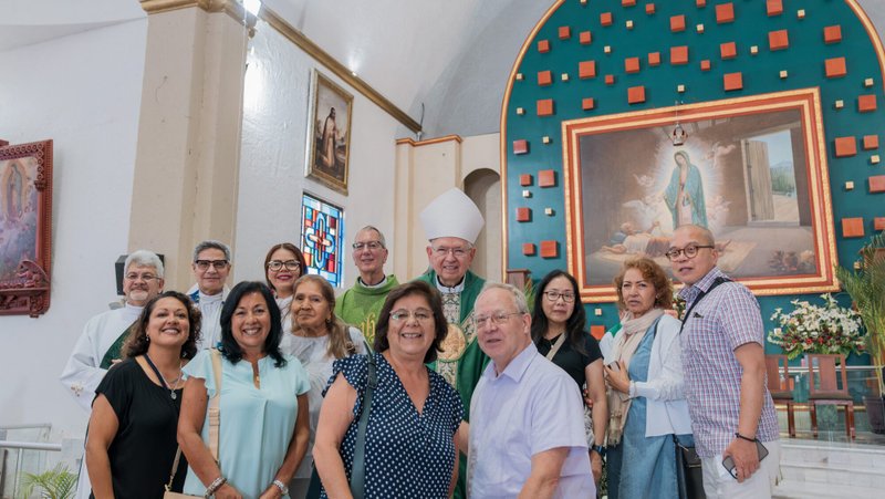 Group of smiling people posing for a photo inside a church with a bishop in green liturgical vestments, near religious artwork and a large teal altar backdrop with decorative squares.