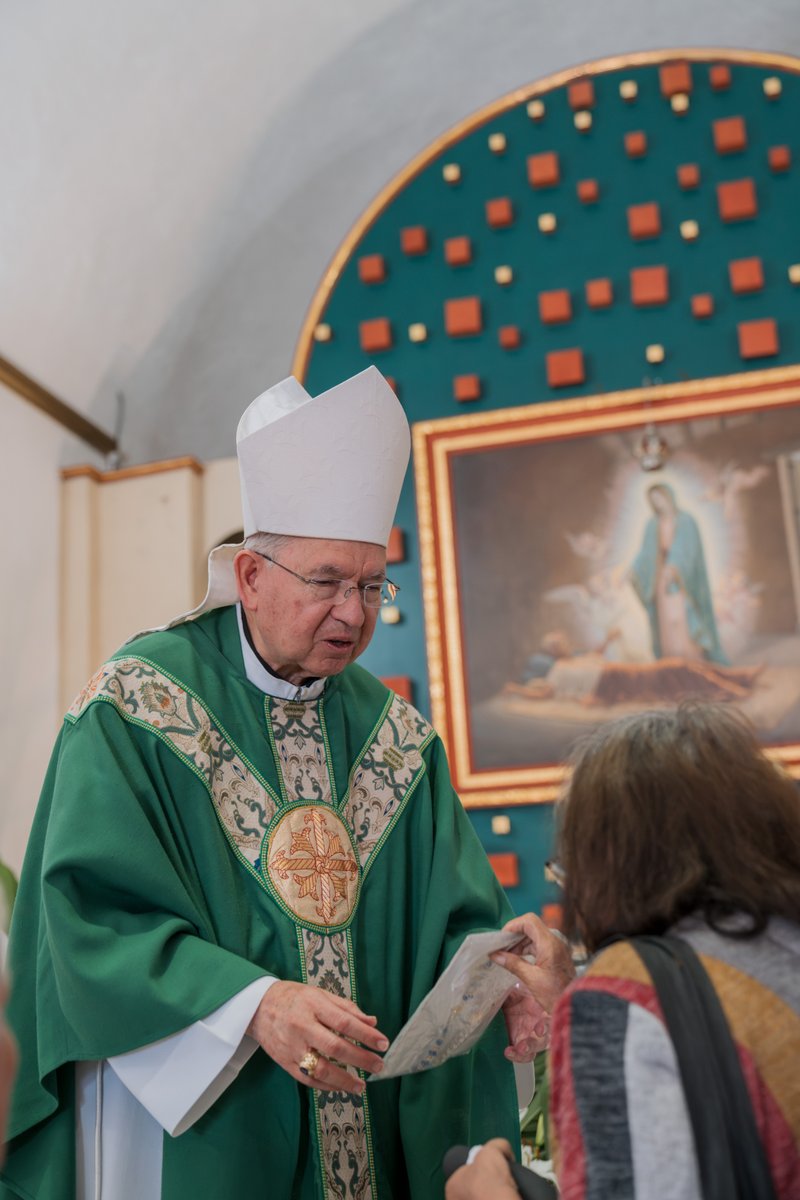 Bishop in green liturgical robes giving a blessing or showing a document to a woman during a church service, with a framed religious painting in the background.