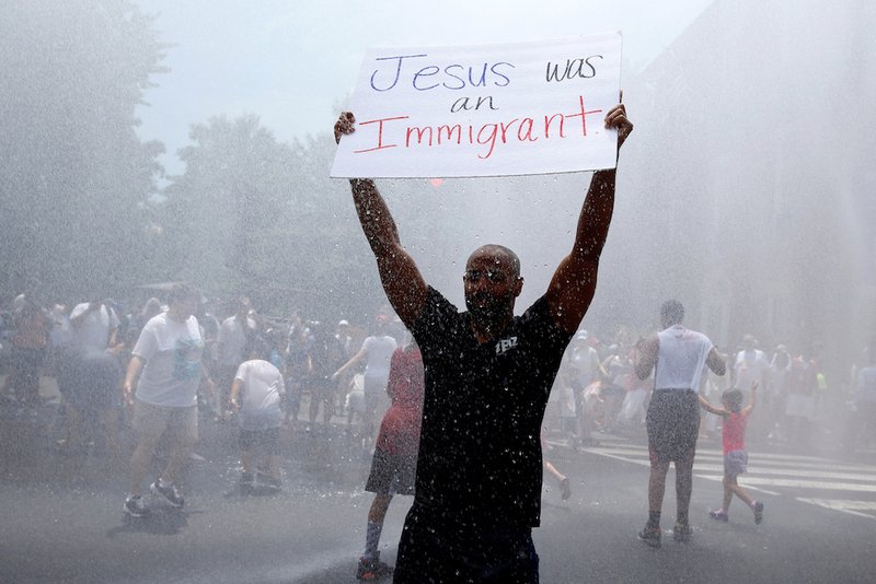 A man holds up a sign as he stands in a spray of water from a fire truck during a late June rally by immigration activists to protest against the Trump administration's immigration policy outside the White House in Washington. Catholic leaders are speaking out against a Trump administration proposed rule announced Sept. 22 that would deny green cards to legal immigrants using public assistance. (CNS photo/Joshua Roberts