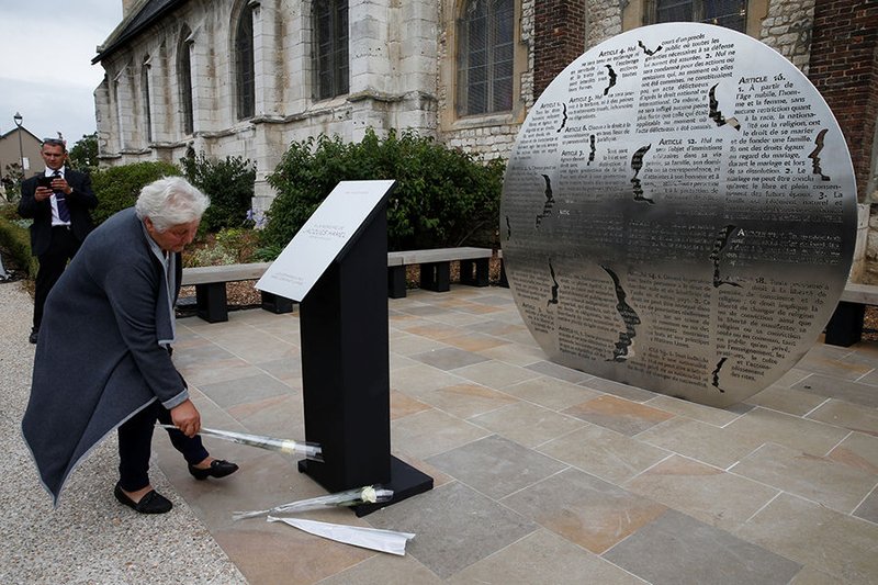 A woman places a flower in front of a plaque and monument in Saint-Étienne-du-Rouvray