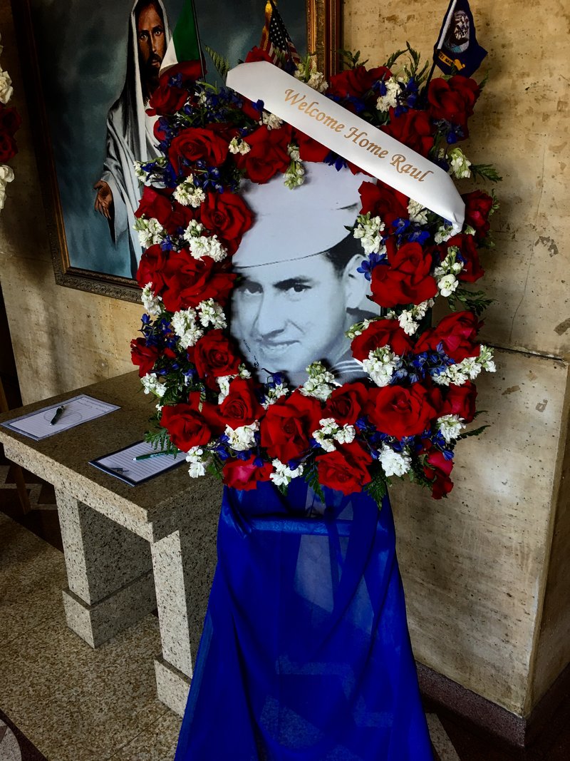 A flower arrangement with Raul Guerra's photo just after he enlisted in the Navy in 1965 is in the vestibule of St. Alphonsus Church for his funeral on April 25. (TOM HOFFARTH)