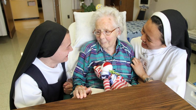 A file photo shows Dominican postulant Anna Harper, a native of Baton Rouge, La., and Dominican Sister Carmela Marie, a native of Hinatuan, Philippines, visit with patient Edna MacCready at Rosary Hill Home in Hawthorne, N.Y. Four orders of women religious and Catholic healthcare ministries filed suit in federal court in Albany July 17, 2026, arguing that New York’s new assisted dying law would force them to choose between their faith and their ability to provide care for the sick, the elderly, and the dying. (OSV News photo/Gregory A. Shemitz)