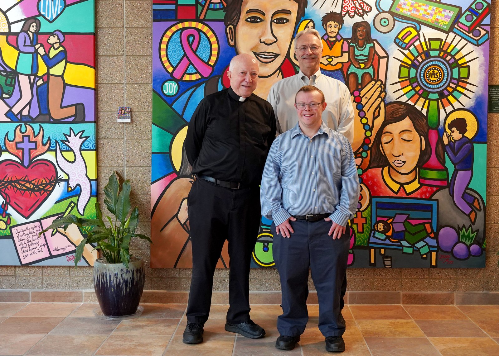 Robby Heil poses with his father, Charles Heil, and Fr. Ed Zaorski, in front of a mural at St. James Parish in Novi. Fr. Zaorski said the Church has made strides in accommodating and welcoming those with disabilities.