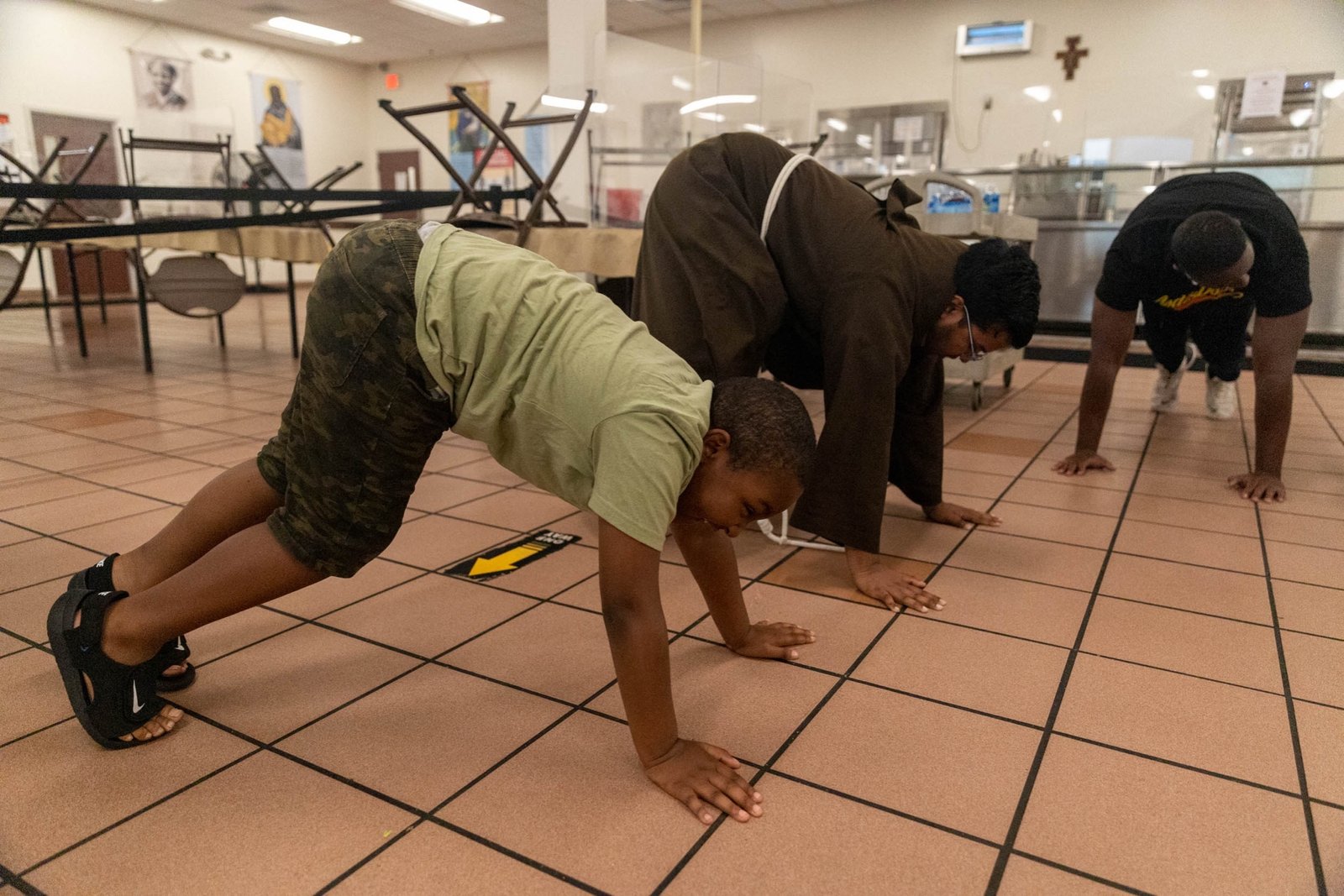 Frailes, campistas y consejeros hacen flexiones durante una actividad de grupo como parte del Campamento de Verano por la Paz Rosa Parks en el Comedor de los Capuchinos. El campamento permite a los niños desarrollar hábitos saludables, practicar la interacción con los demás y divertirse.